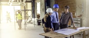 Three people standing at a table with stretched out plans on it and talking about the construction process and costs.