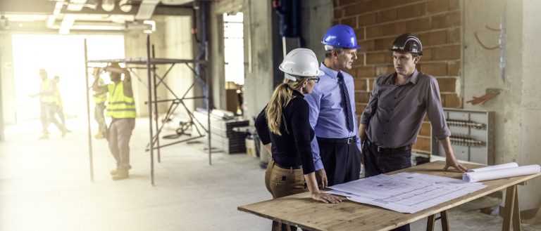 Three people standing at a table with stretched out plans on it and talking about the construction process and costs.